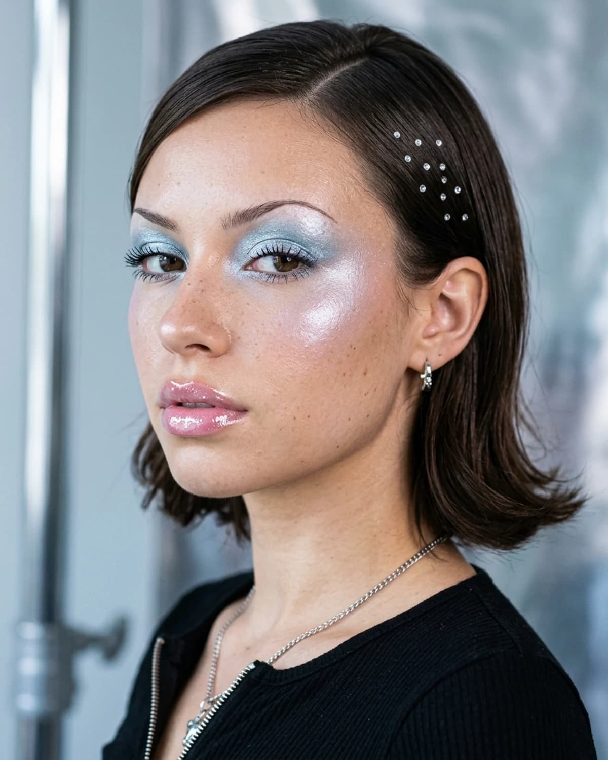 A woman wearing a Y2K makeup look: frosted icy blue eyeshadow, thin arched brows, shimmery cheeks and super-glossy pink lips.